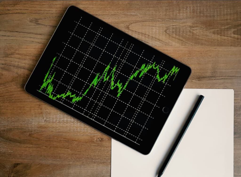 Tablet with stock market data graph displayed on screen, resting on a wooden table next to a stylus and a blank sheet of paper