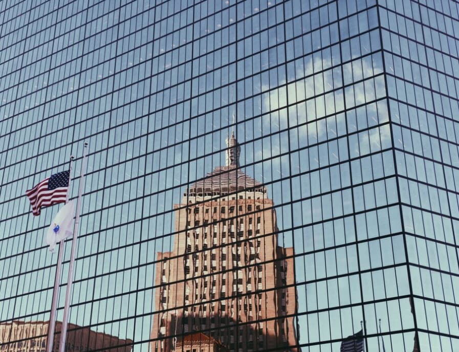 A reflective glass skyscraper mirroring an older building under a clear sky
